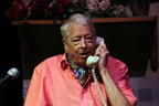 An older man wearing a pink shirt and ascot on the phone