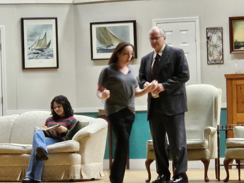 A girl sitting on the couch reading, and a standing woman talking to a standing man in a dark gray suit.