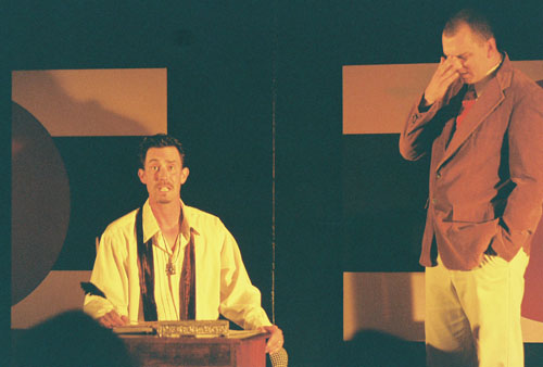 A man at a desk talking to a man in a coat standing
