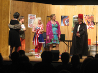 A woman in a black dress and mink stole, a man in a sparkling gold shirt, a woman in a red, white and blue blouse and dress, a woman in a
       sparkling purple coat, and a man in a suit, black cloak and red turban