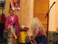 A woman in a sparkling purple coat sitting, talking to a woman in a red, white and blue blouse and skirt