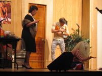 A woman in a black dress and mink stole, a man in zebra-striped pants holding a dog, and a woman in a red, white and blue blouse and skirt kneeling