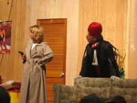 A woman in a raincoat holding a gun and a man in a suit, black robe and red turban next to her