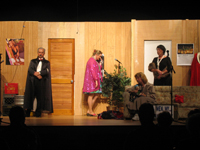 A man in a suit and black cloak, a woman in a sparkling purple coat, a woman in a raincoat sitting on a couch and a woman in a black dress and mink stole standing