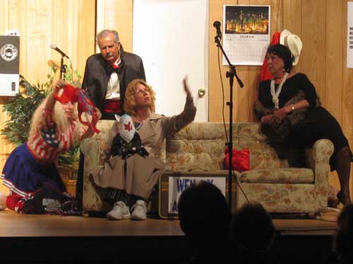 A woman in a red, white and blue dress kneeling next to a couch on which a woman in a brown raincoat and a woman in a black
       dress sit, with a man in a suit standing behind the couch 