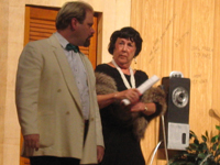A man in a white tuxedo jacket standing next to a woman in a black dress and mink stole
