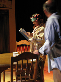A woman in a robe standing and reading a magazine while a man stands next to a table