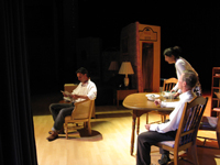 A man sitting in a chair, another man sitting at a table and a standing woman talking