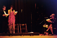 A woman in a slit skirt and a man standing at a microphone, and a woman sitting at a microphone