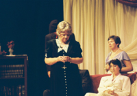 An older woman with a drink, a sad woman on a couch and another woman behind them