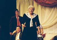 A woman with a handbag standing in front of a sitting man in a suit and a man in a beret