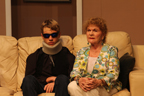 On a couch, a woman wearing a print jacket talking to a man wearing a neckbrace and sunglasses