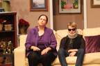 On a couch, a woman wearing a purple outfit talking to a man wearing a neckbrace and sunglasses