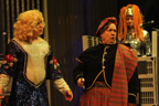 The man in the wig and white and blue skirt stands next to a woman wearing a plaid outfit and beret