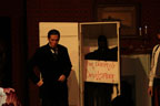 A man in a dark suit standing fron front of a tall box with a sign that says The Doom of Devilsmoor