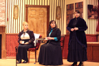 A woman in a white blouse and dark jacket and the woman in a dark dress and blue blouse sit while a woman in a long dark
       dress talks 