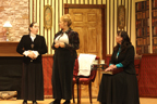 A woman in a black blouse talks to the woman in the white blouse and dark jacket while the woman in the dark dress and blue
       blouse sits and listens