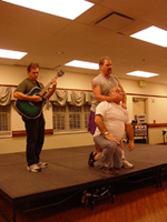 A man holding a man's mouth shut while a third man plays guitar