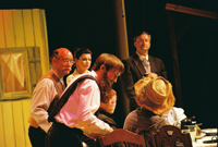 A man in a beard talks to a seated man in a hat while two men and two woman look on