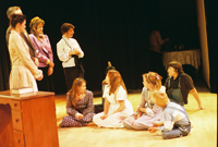 A woman talking to six children sitting on the floor with two women and one man looking on