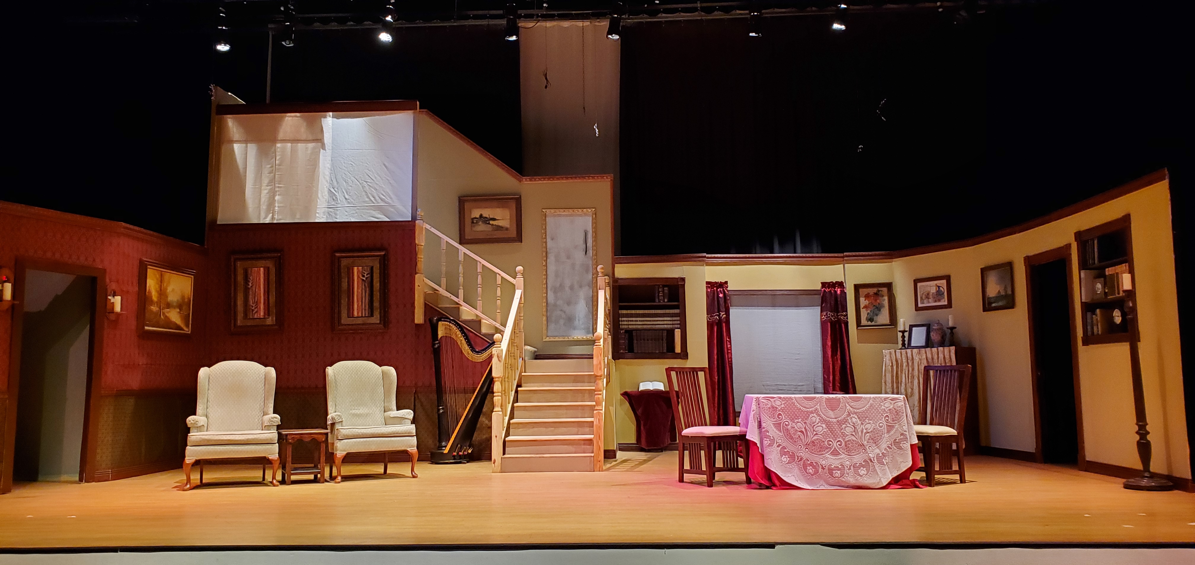 A theater set with reddish brown walls on the left and light brown walls on the right, and a set of stairs just to the left of center. The stairs turn
       left at a landing and go up again. A harp sits near the stairs. Two beige chairs sit to the left and a table and two chairs sit toward the right. There is a
       large window with red drapes toward the back on the right. There are doorways to the left and right.