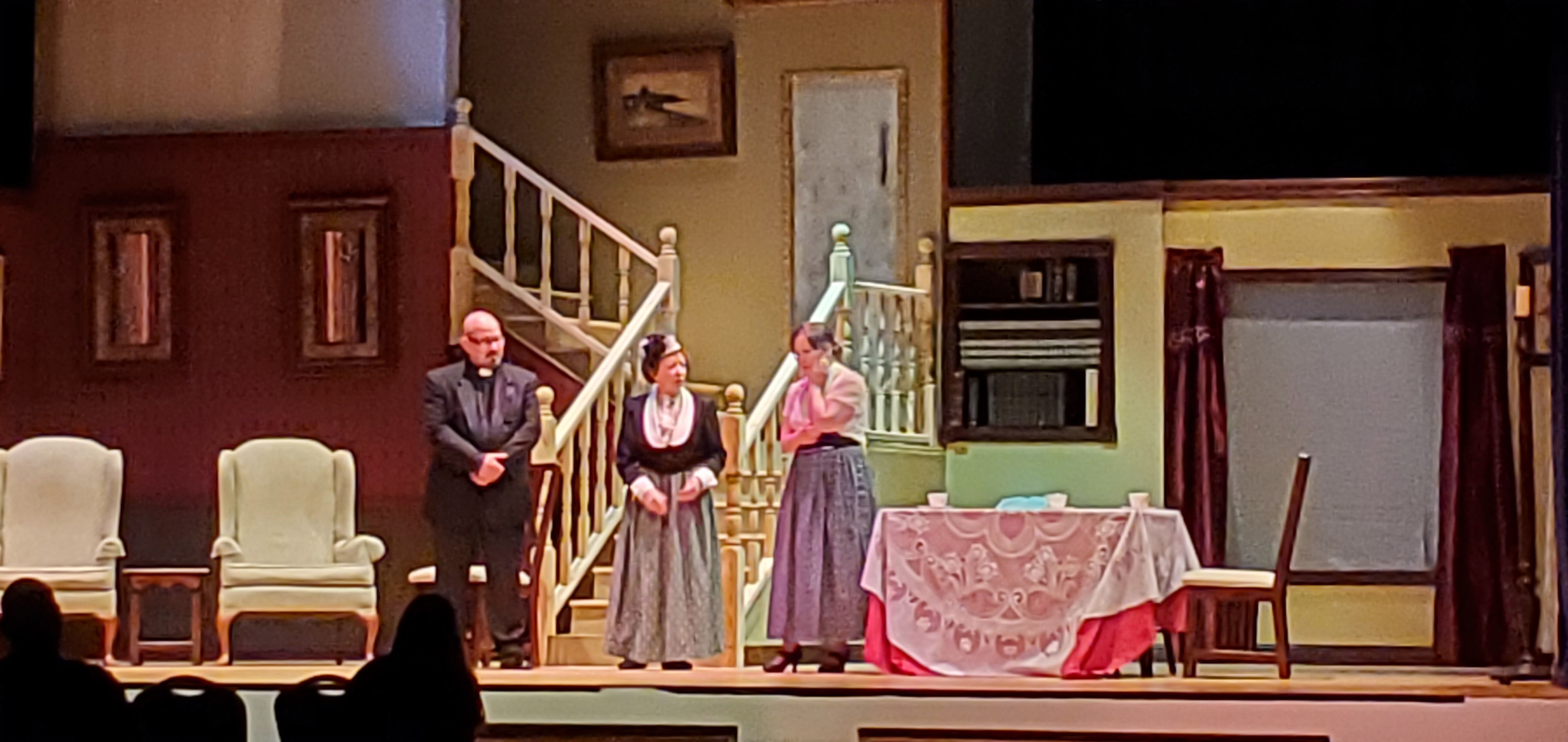 A priest, a woman in Victorian dress and another woman in a white blouse and a long gray dress stand near the steps. A table with a white tablecloth
       and and rose-colored cloth underneath is to the right of the people.