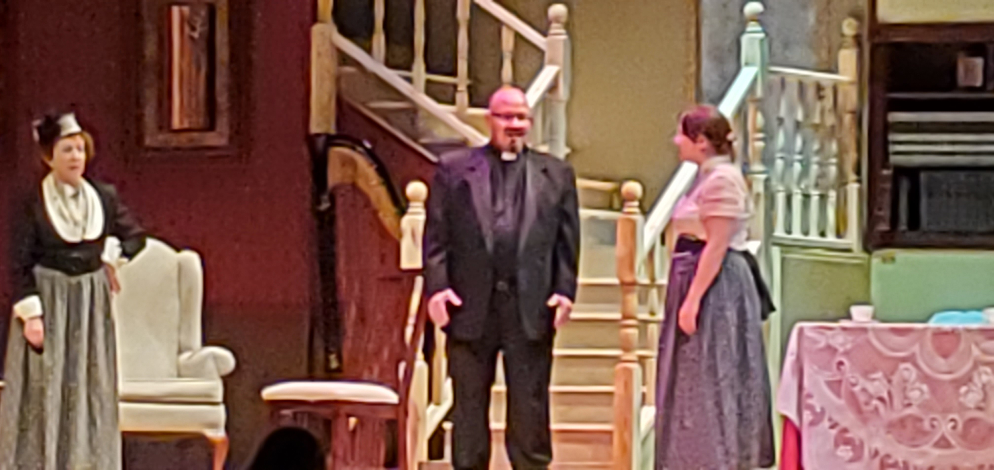 A woman in Victorian dress is to the left, the priest is in the middle and another woman in a white blouse and a long gray dress stands to the right.
       They stand in front of a staircase.