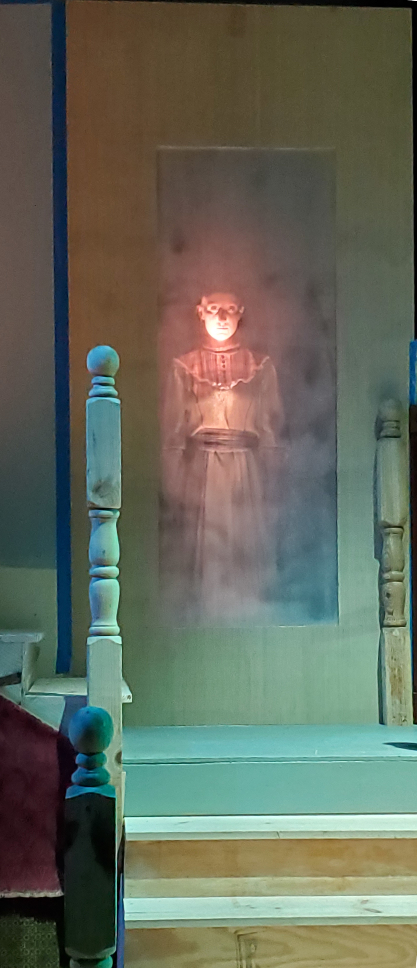 A girl standing behind a scrim on the first landing of the stairway in eerie light and surrounded by smoke.