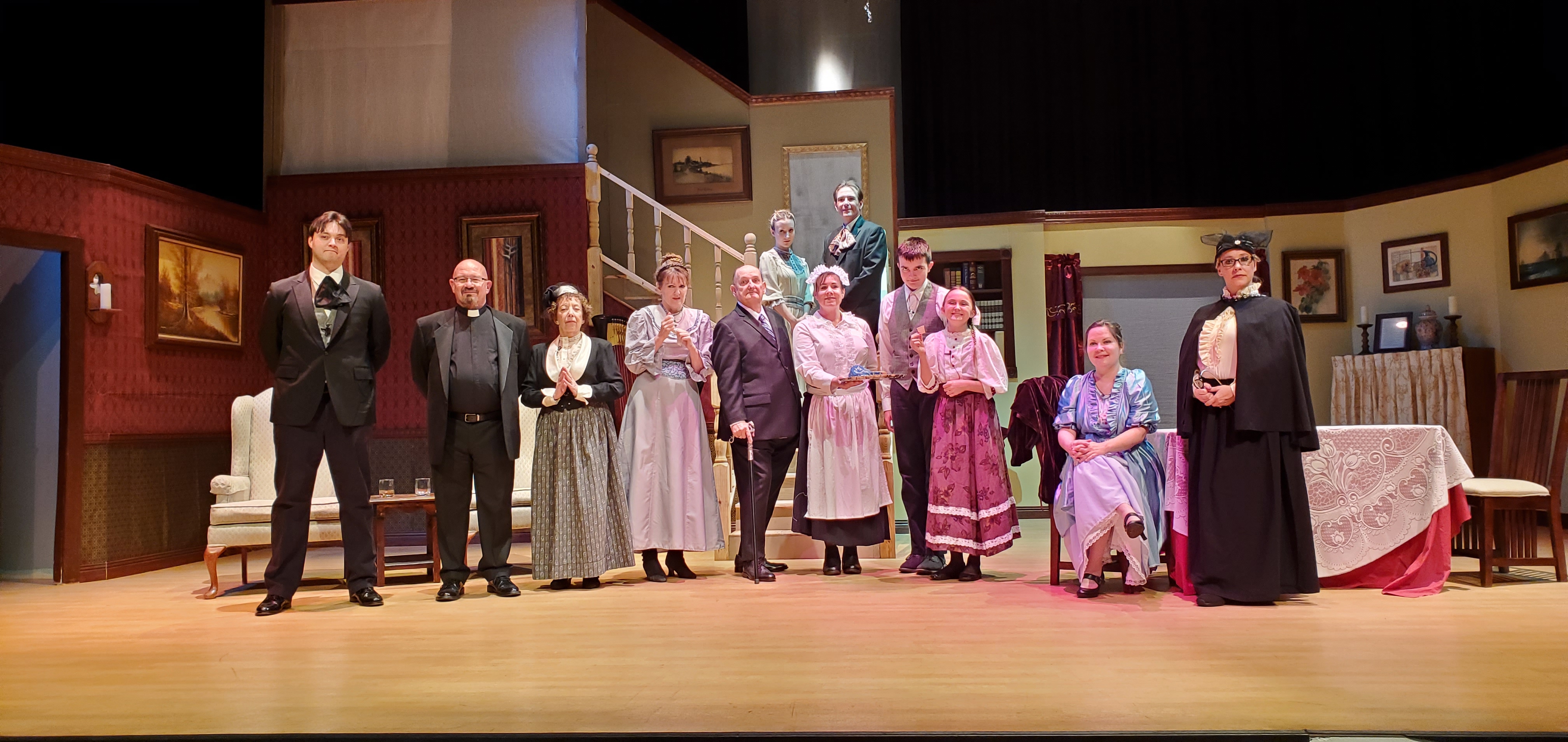 The entire cast standing, from left to right: a man in a black suit and ascot, a priest, a woman in Victorian dress, a woman in a long gray dress
       a man in a suit holding a cane, a maid in a white outfit, a tall boy in a white shirt and gray vest, a girl in a light rose top and rose colored dress, a woman in a metallic blue
       colored dress sitting, a woman in a black cloak and black dress standing. A teenage girl in a light green dress and a teenage boy in a gray suit stand behind and up on the
       landing.