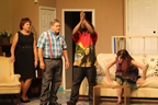 A woman in a black dress, a man wearing a blue plaid shirt and red suspenders, a man wearing a Bob Marley shirt, and a woman getting up off the couch