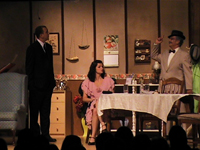 Man in suit and man in suit and hat standing, with one woman sitting at a table